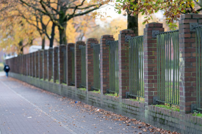 Eine Mauer aus Steinsäulen und Gittern an einem Fußweg, grenzt einen Platz von der Straße ab