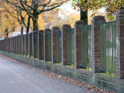 Eine Mauer aus Steinsäulen und Gittern an einem Fußweg, grenzt einen Platz von der Straße ab